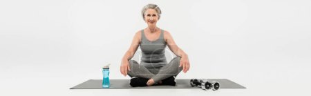 longitud completa de la mujer mayor feliz sentada en postura del yoga en la estera cerca de las mancuernas y de la botella del deporte en gris, bandera 