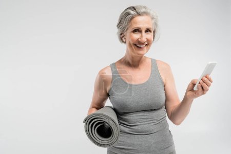 Photo for Carefree senior woman in wireless earphones holding fitness mat and smartphone isolated on grey - Royalty Free Image