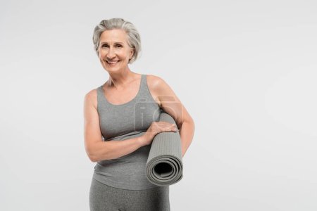 Foto de Alegre sénior mujer en ropa deportiva celebración de fitness mat y sonriendo aislado en gris - Imagen libre de derechos