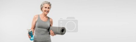 Foto de Alegre anciana sosteniendo alfombra de fitness y botella de deporte con agua aislada en gris, pancarta - Imagen libre de derechos
