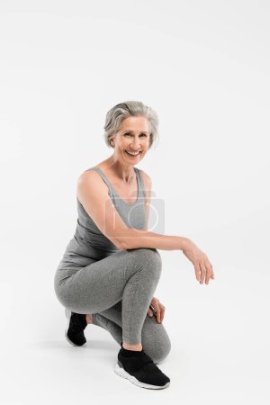 Foto de Longitud completa de la alegre mujer mayor en ropa deportiva sonriendo mientras está sentado en gris - Imagen libre de derechos