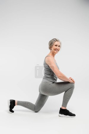 Foto de Longitud completa de la mujer mayor feliz en ropa deportiva haciendo saltos y sonriendo en gris - Imagen libre de derechos
