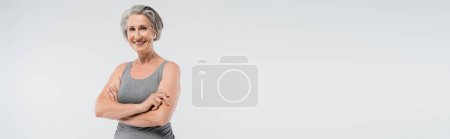 carefree senior woman in sportswear standing with folded arms isolated on grey, banner 
