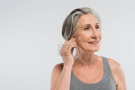 happy senior woman adjusting wireless earphone isolated on grey 