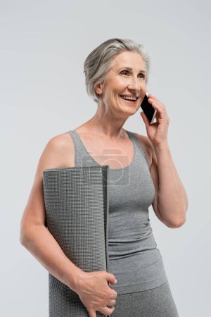 happy grandma holding fitness mat and talking on mobile phone isolated on grey 