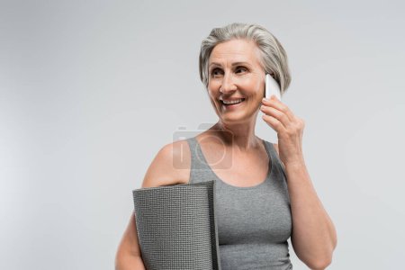 Photo for Overjoyed grandma holding fitness mat and talking on mobile phone isolated on grey - Royalty Free Image