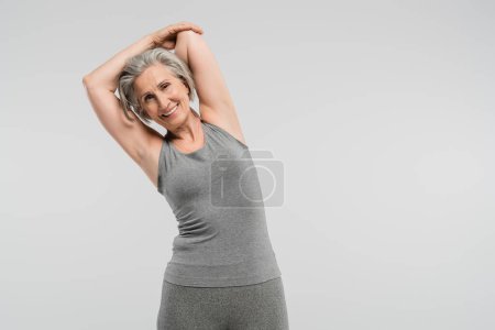 Foto de Alegre abuela en ropa deportiva ejercitando y sonriendo aislado en gris - Imagen libre de derechos