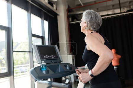 retired woman with grey hair running on treadmill next to sports bottle with water 