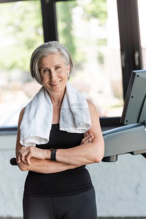 positive senior woman with white towel on shoulders standing next to treadmill 