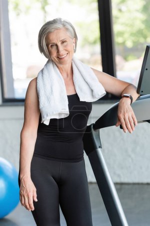 joyful senior woman with white towel on shoulders standing next to treadmill 