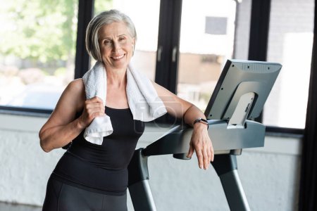joyful retired woman with white towel on shoulders standing next to treadmill 