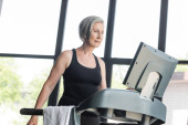 retired woman with grey hair looking at monitor of treadmill while working out in gym  mug #648885148