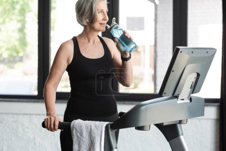 Foto de Mujer mayor con el pelo gris beber agua de la botella de deportes después de ejercicio cardiovascular en la cinta de correr - Imagen libre de derechos