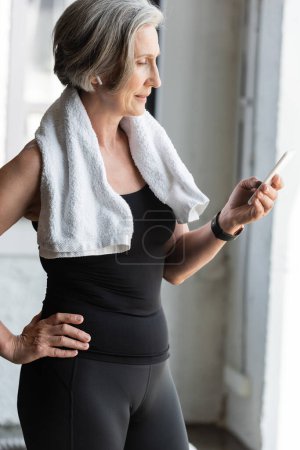 fit senior woman with towel on shoulders using smartphone while listening music in wireless earphone 