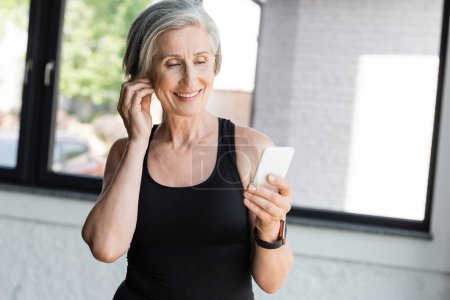 fitte Seniorin in schwarzem Tanktop mit Smartphone beim Musikhören im drahtlosen Kopfhörer 