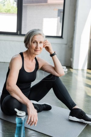 mujer mayor en ropa deportiva escuchar música y sentarse en la alfombra de fitness en el gimnasio 