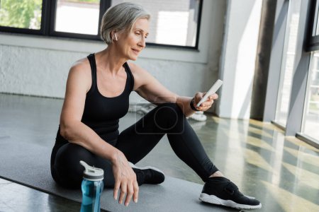 Téléchargez les photos : Femme âgée en vêtements de sport en utilisant un téléphone portable et assis sur tapis de fitness dans la salle de gym - en image libre de droit