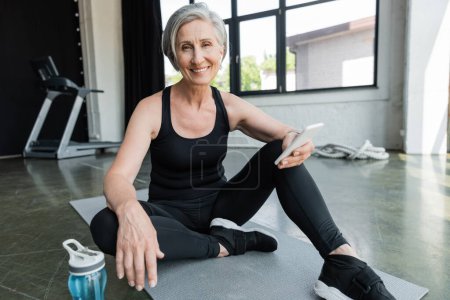 fröhliche Sportlerin mit grauen Haaren nutzt Handy und sitzt auf Fitnessmatte im Fitnessstudio 