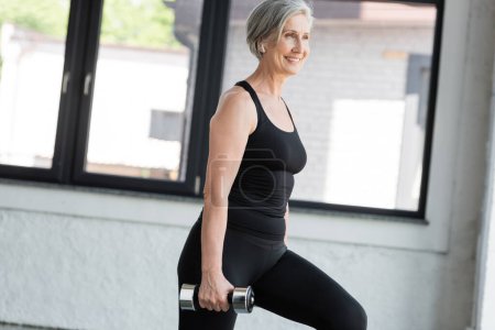 positive senior woman in wireless earphone listening music while working out with dumbbell in gym 
