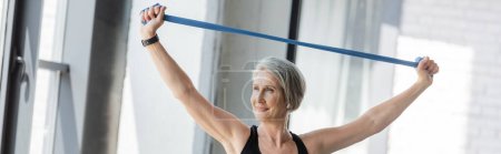 happy senior woman working out with resistance band above head in sports center, banner 