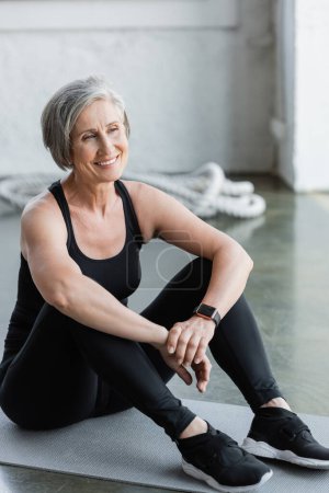 happy senior woman in black leggings and tank top sitting on fitness mat 