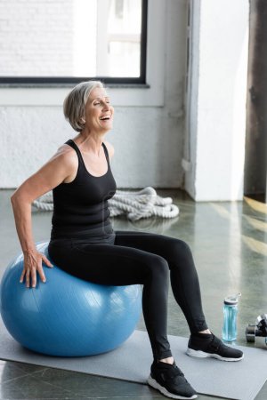 full length of excited senior woman in black leggings and tank top exercising on blue fitness ball 