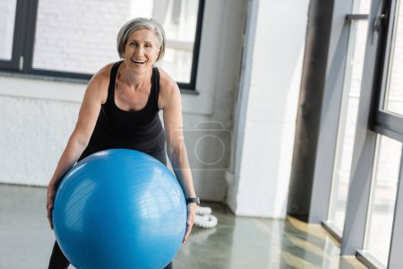 Téléchargez les photos : Excitée femme âgée en vêtements de sport noirs tenant ballon de remise en forme bleu tout en travaillant dans la salle de gym - en image libre de droit