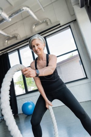 cheerful senior sportswoman with grey hair working out with battle ropes in gym 