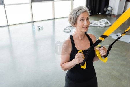 Hochwinkelaufnahme einer fitten Seniorin in schwarzer Sportbekleidung, die mit Federbeinen im Fitnessstudio trainiert 