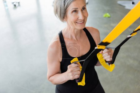 Blick aus der Vogelperspektive auf eine fröhliche Seniorin in schwarzer Sportbekleidung, die im Fitnessstudio mit Federbeinen trainiert 