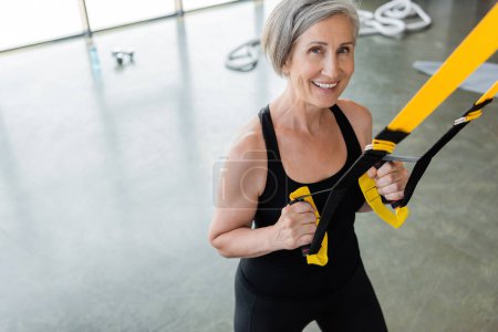 Blick aus der Vogelperspektive auf eine glückliche Seniorin in schwarzer Sportbekleidung, die mit Federbeinen im Fitnessstudio trainiert 