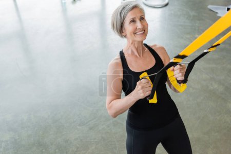 Blick aus der Vogelperspektive auf eine überglückliche Seniorin in schwarzer Sportbekleidung, die mit Federbeinen im Fitnessstudio trainiert 