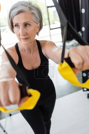 Téléchargez les photos : Senior femme en noir sportswear exercice avec des bretelles de suspension au premier plan floue - en image libre de droit