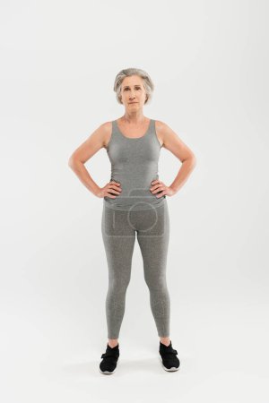 Foto de Longitud completa de la mujer feliz y jubilada en ropa deportiva de pie con las manos en las caderas en gris - Imagen libre de derechos