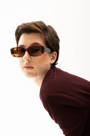brunette woman in trendy sunglasses and brown blazer looking at camera isolated on white