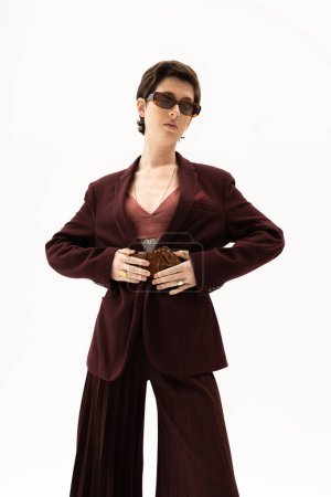 modelo morena en traje de pantalón marrón burdeos y gafas de sol tocando cinturón de cuero aislado en blanco