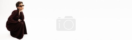Téléchargez les photos : Pleine longueur de femme avec des cheveux bruns courts posant en costume marron et des lunettes de soleil élégantes sur fond blanc, bannière - en image libre de droit