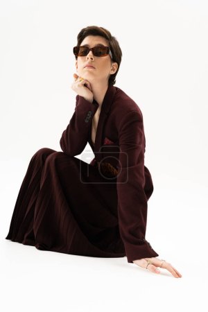 full length of young woman in stylish suit and sunglasses posing on haunches on white background