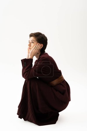 dreamy woman in brown suit holding hands near face and looking away on white background