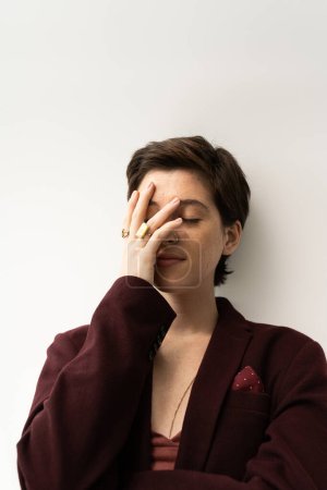 brunette model in brown jacket and finger rings obscuring face with hand on grey background