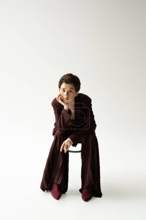 full length of woman with short brunette hair sitting on chair in elegant pantsuit and looking at camera on grey background