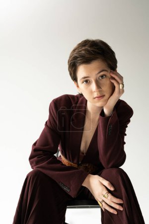 brunette model in brown suit and finger rings sitting and looking at camera on grey background