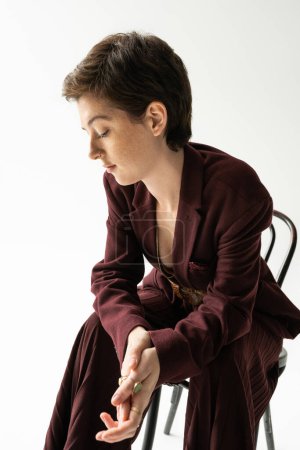 dreamy woman in elegant pantsuit looking down while sitting on chair on grey background