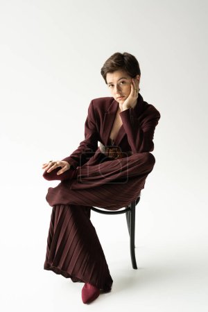 young and elegant woman in wide pants looking at camera while posing on chair on grey background