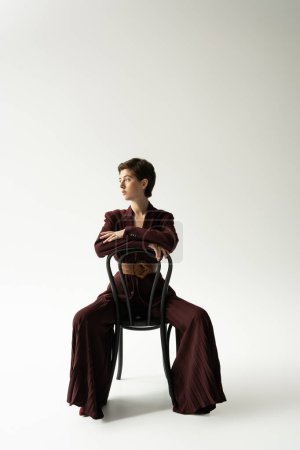 full length of brunette model in blazer and wide pants sitting on chair and looking away on grey background