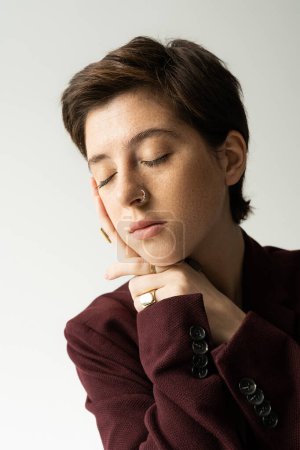 portrait of young freckled woman in brown blazer posing with closed eyes isolated on grey