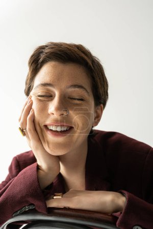 Foto de Retrato de mujer joven alegre en blazer de moda y anillos de dedo sonriendo con los ojos cerrados aislados en gris - Imagen libre de derechos