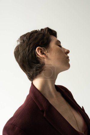 Téléchargez les photos : Vue latérale de la jeune femme sensuelle en boucle d'oreille manchette et blazer élégant isolé sur gris - en image libre de droit