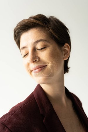 Porträt einer glücklichen jungen Frau mit kurzen brünetten Haaren, die mit geschlossenen Augen lächelt, isoliert auf grau
