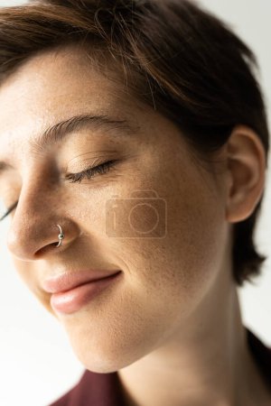Nahaufnahme Porträt einer unbeschwerten Frau mit Sommersprossen und Nasenpiercing lächelnd mit geschlossenen Augen auf grauem Hintergrund